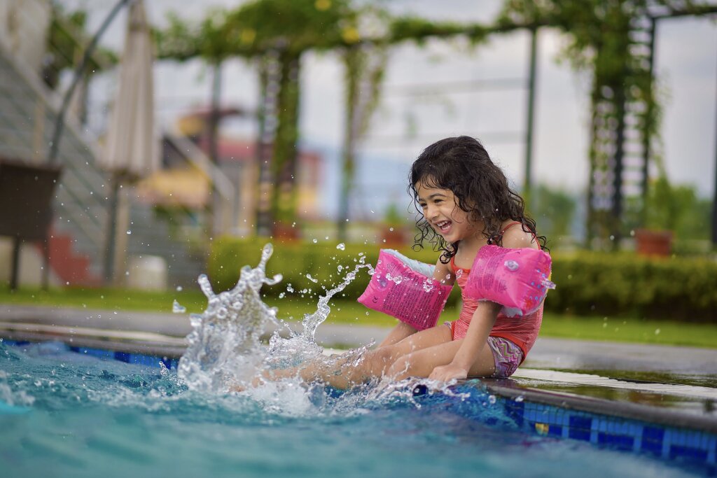 Een kind met roze zwembandjes lachend op de rand van een zwembad trappelend in het water.