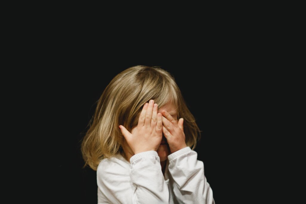 Een kindje voor een zwarte achtergrond dat schuilt achter haar handen.