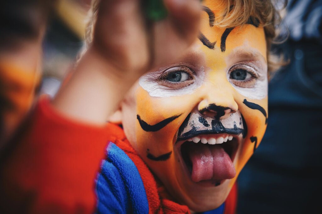 Een blij kind in een superheldpakje met een geschilderd gezicht. Kijkt lachend in de camera met mond wijd open.