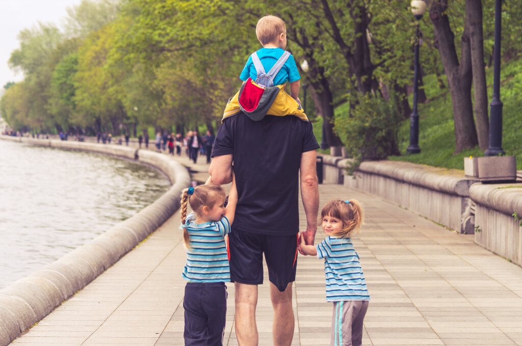 Een vader van achteren gefotografeerd met twee kinderen aan de hand en één kind op de schouders.
